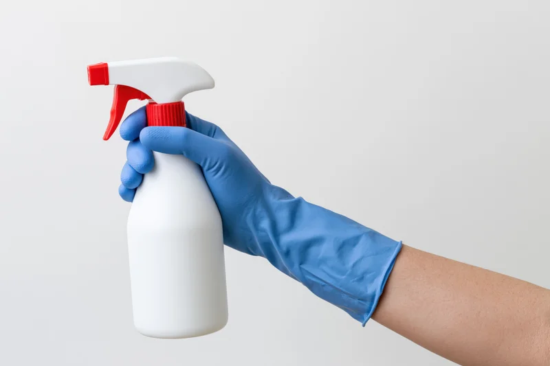 Hand in blue cleaning glove holding white spray bottle with red nozzle against plain background