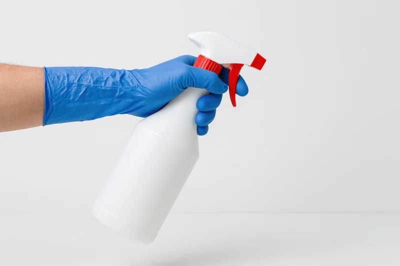 Hand in blue rubber glove holding white spray bottle against plain white background