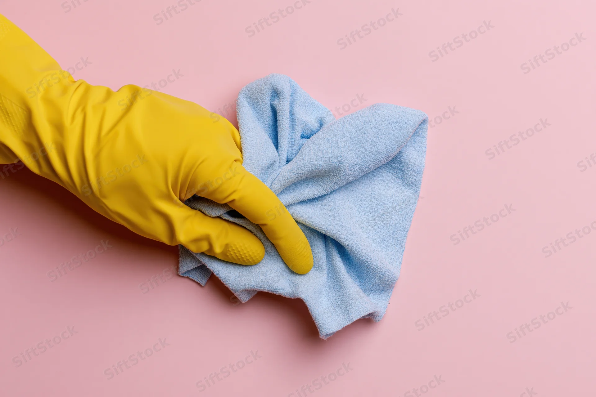 Hand in yellow rubber glove holding blue cloth on pink surface