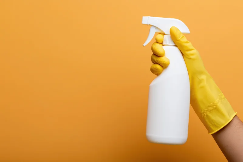Hand in yellow rubber glove holding white spray bottle against orange background