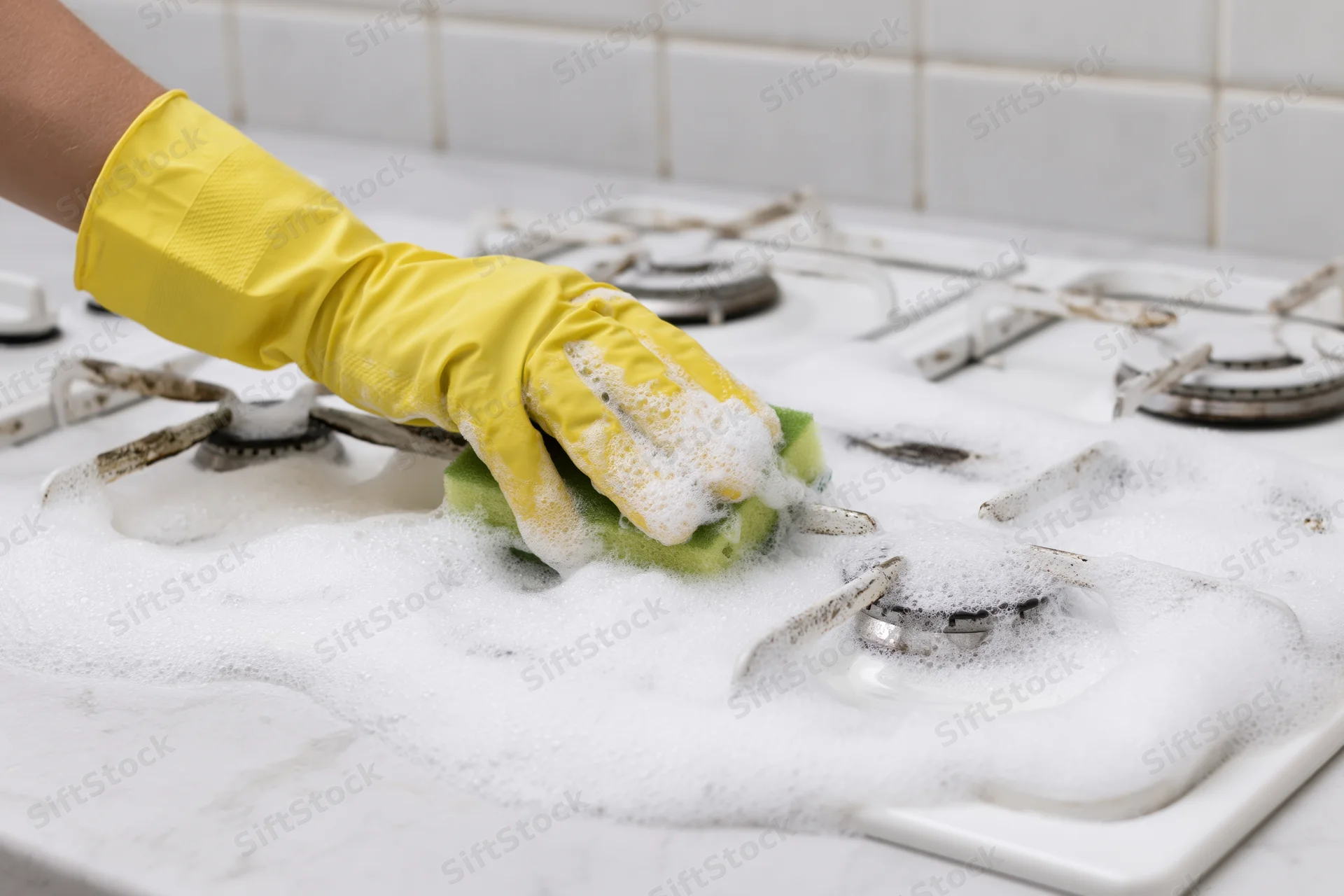 Hand in yellow rubber glove scrubbing soapy white gas stove with green sponge
