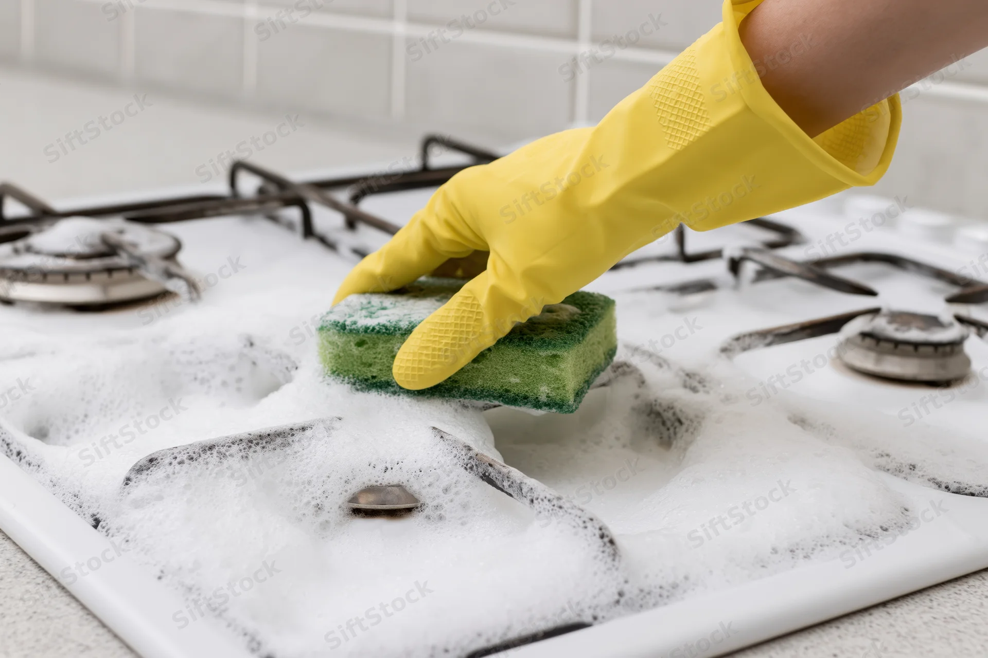 Hand in yellow rubber glove scrubbing soapy white gas stove with green sponge