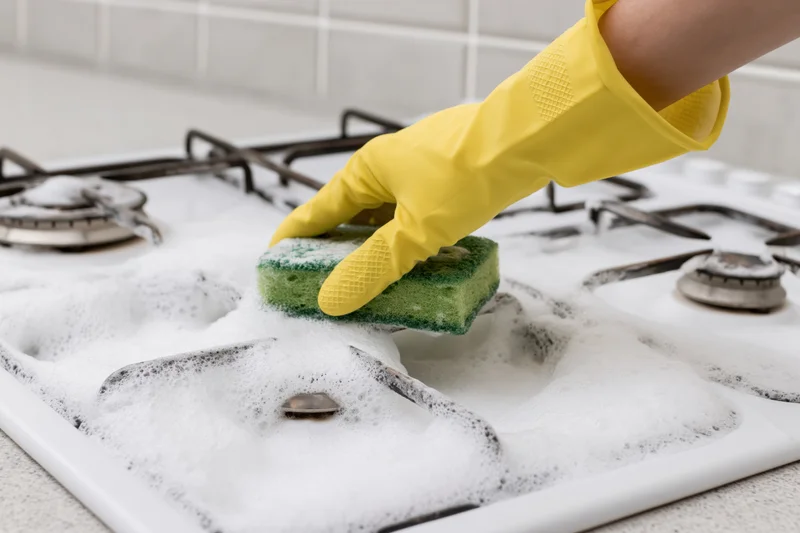 Hand in yellow rubber glove scrubbing soapy white gas stove with green sponge