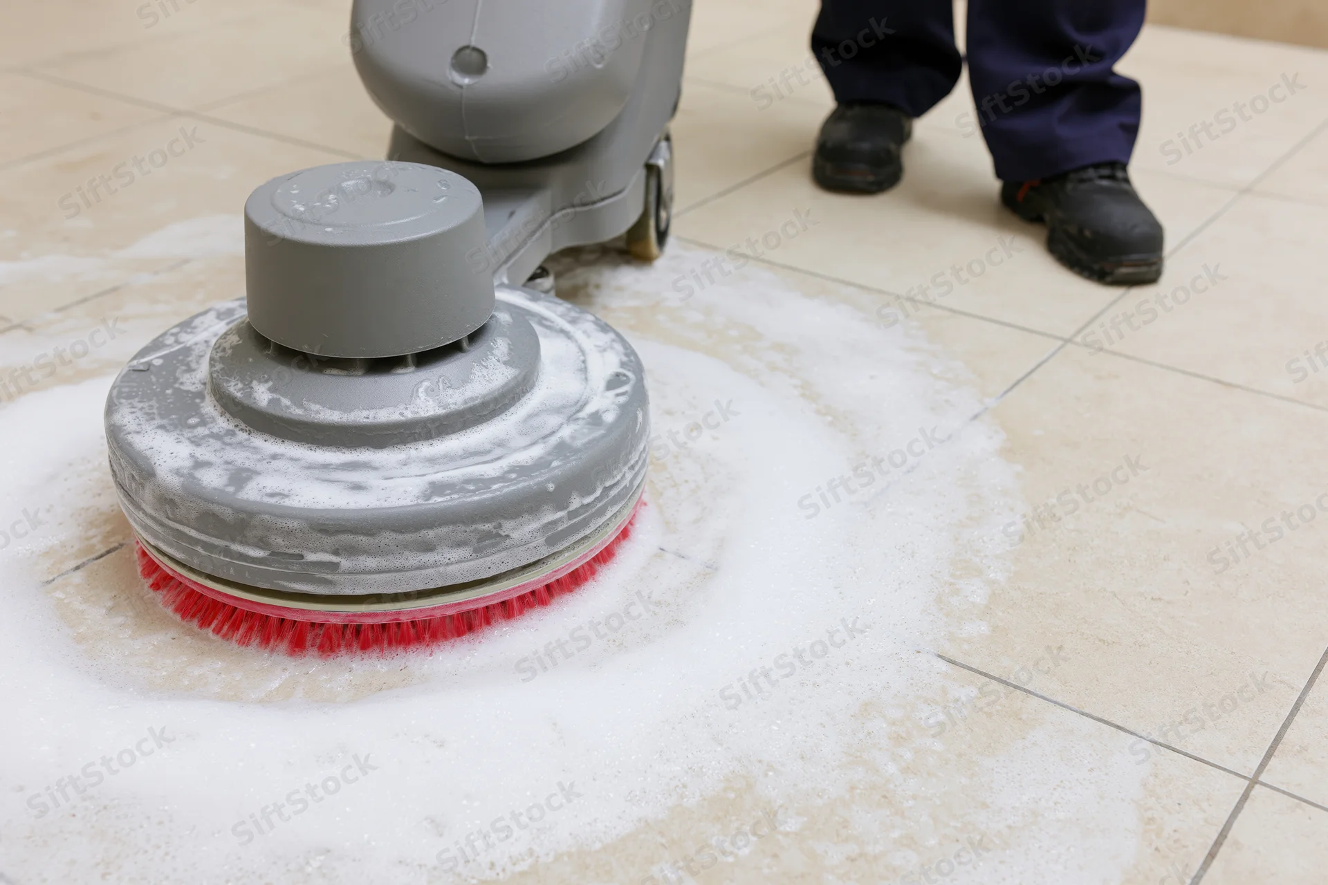 Janitor operating a grey rotary floor scrubbing machine on a tiled floor with soapy foam