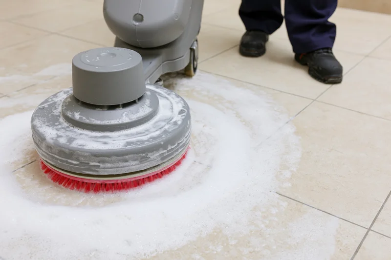 Janitor operating a grey rotary floor scrubbing machine on a tiled floor with soapy foam