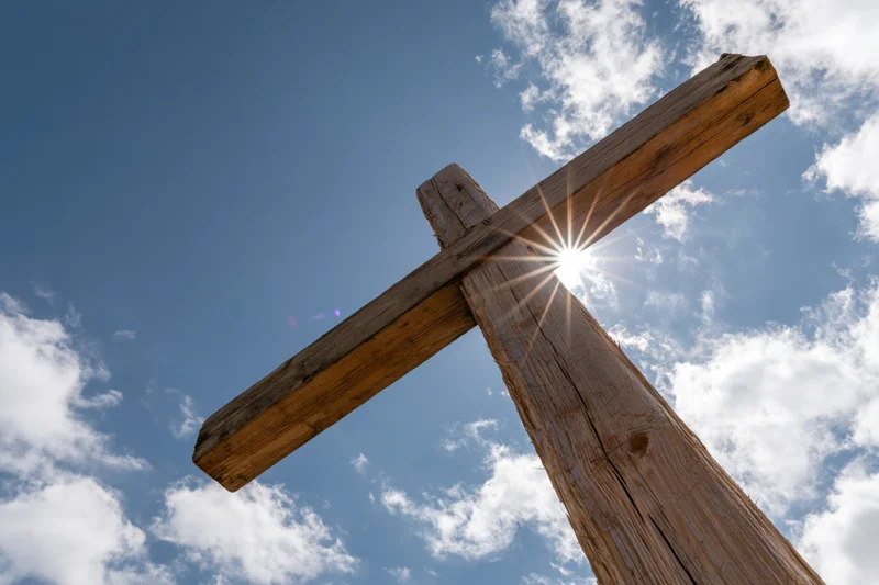 Weathered wooden christian cross against blue sky with sun shining through