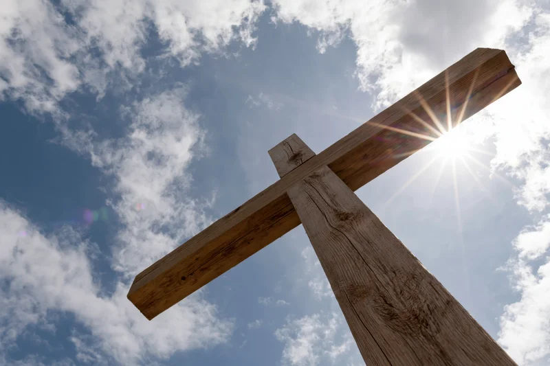 Wooden christian cross against a blue sky with sun shining through clouds