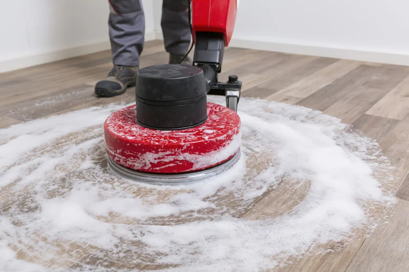 Maintenance worker operating red and black rotary floor polisher on laminate flooring with white foam, interior room