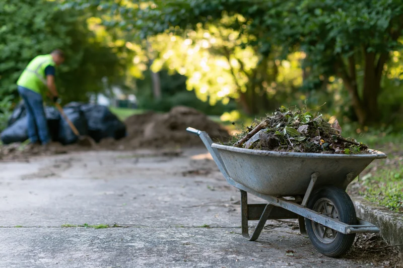 Metal wheelbarrow filled with garden debris on pavement near a worker bagging waste in a park