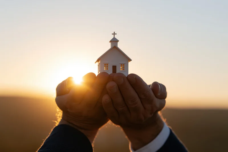 Hands cupping a small white church model against a sunset sky