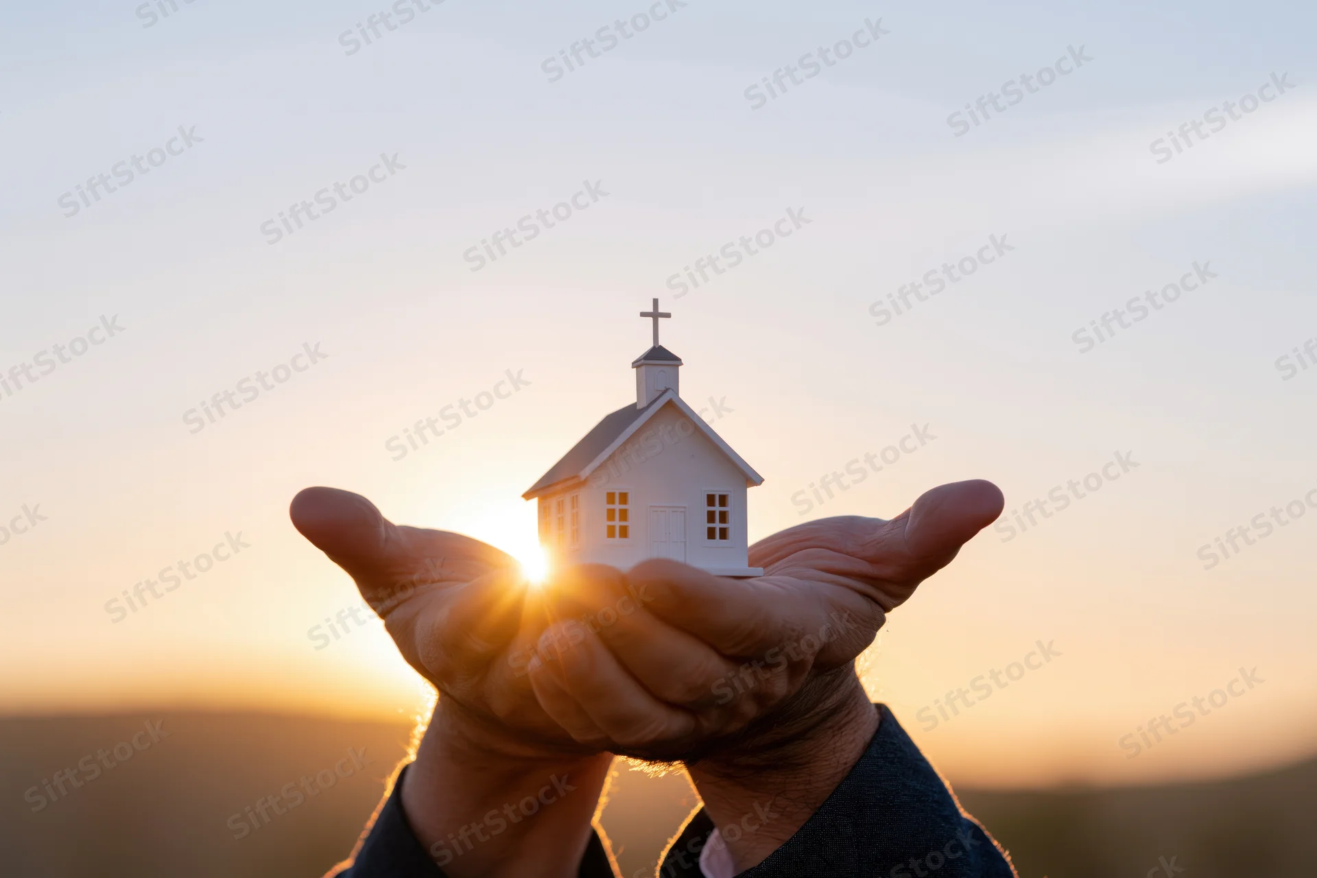 Hands cupping a small white church model against a sunset sky