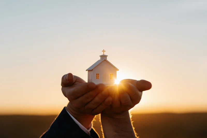 Hands cupping a small white church model against a sunset sky