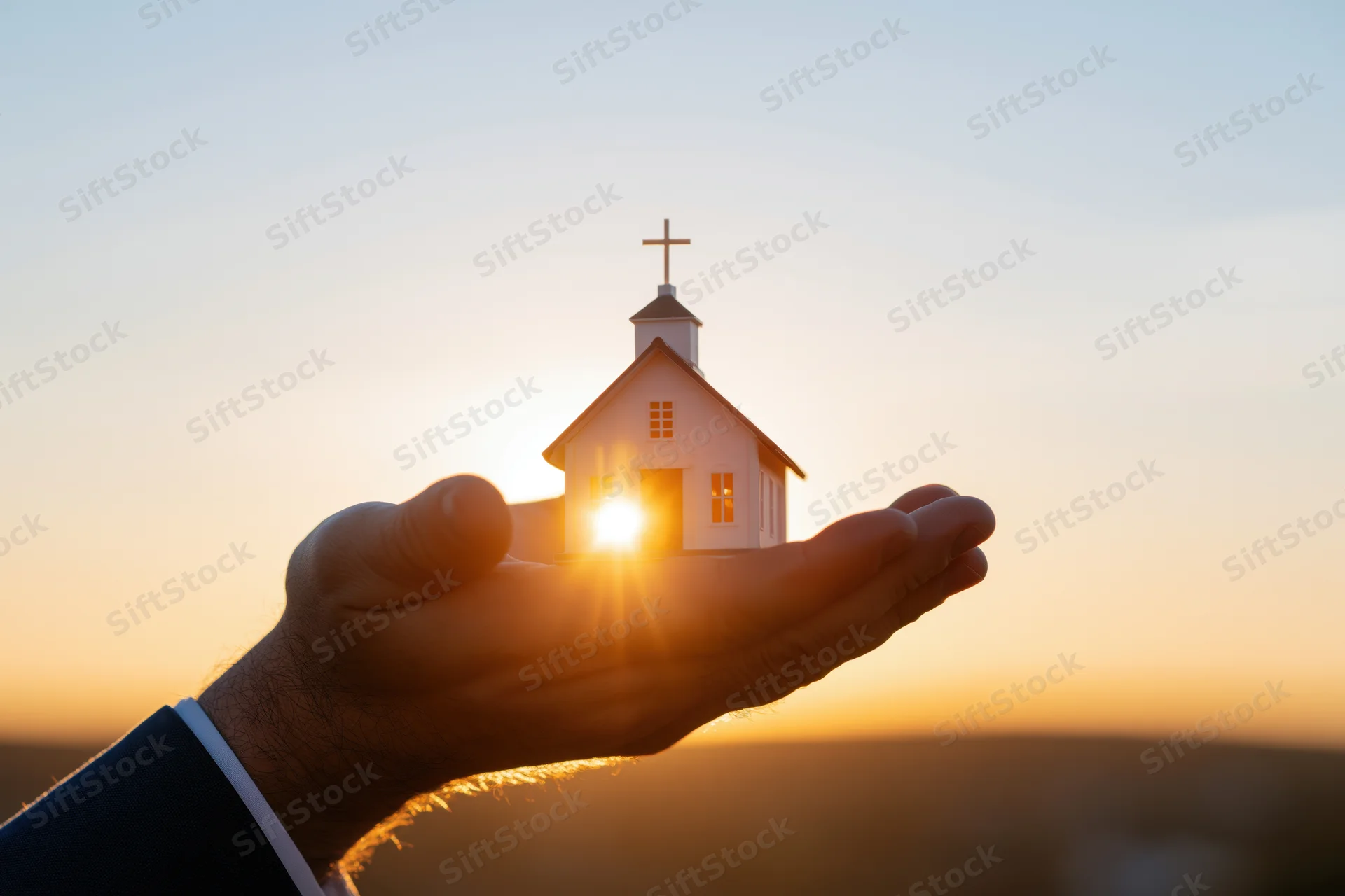 Hands cupping a small white church model against a sunset sky