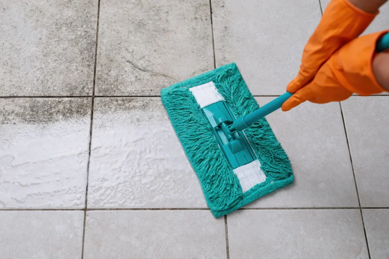Hand in orange rubber glove pushing teal flat mop across tiled floor