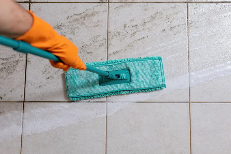 Hand in orange glove pushing teal flat mop across wet tiled floor