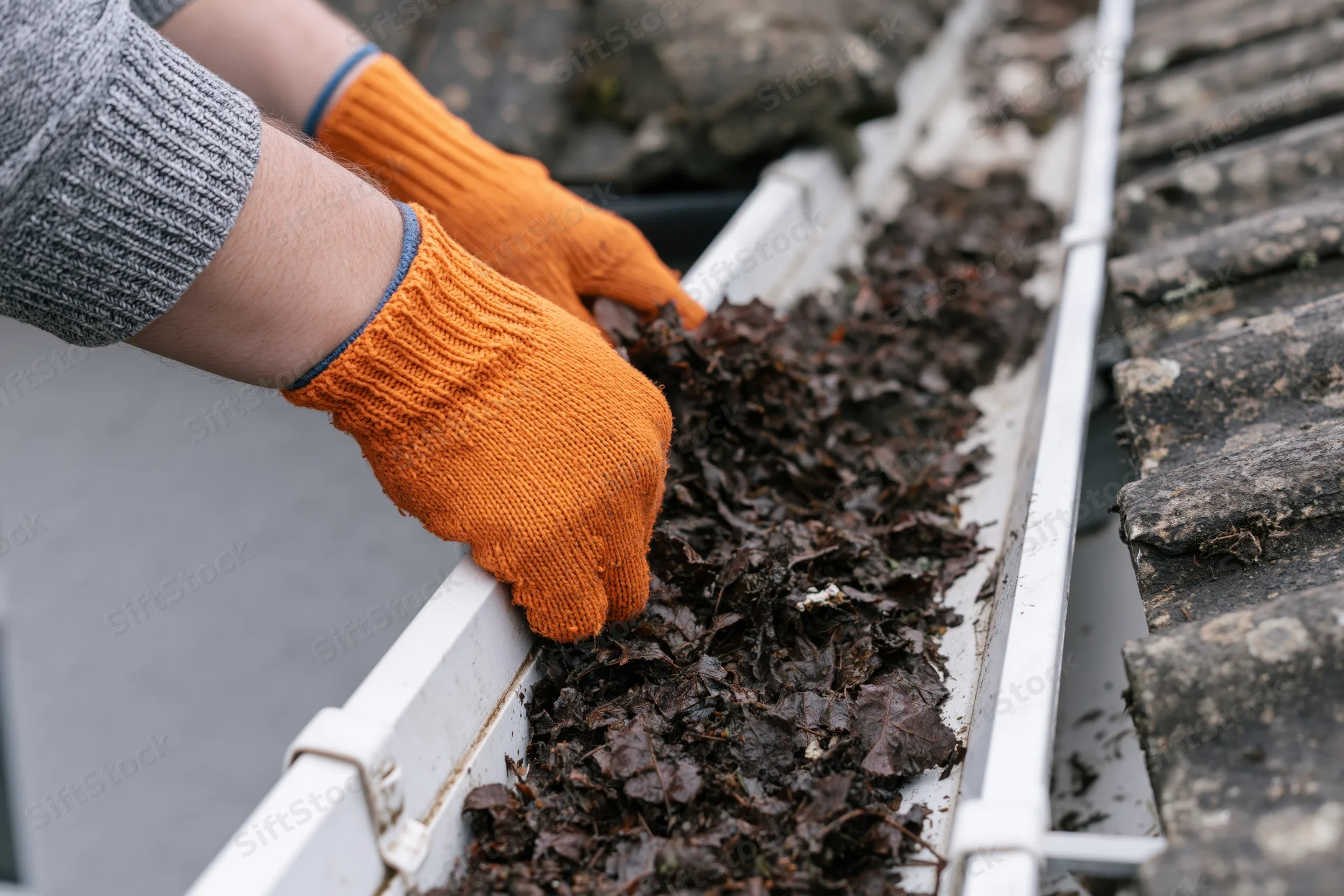 Hands in orange knit gloves clearing wet leaves from a white gutter on a tiled roof