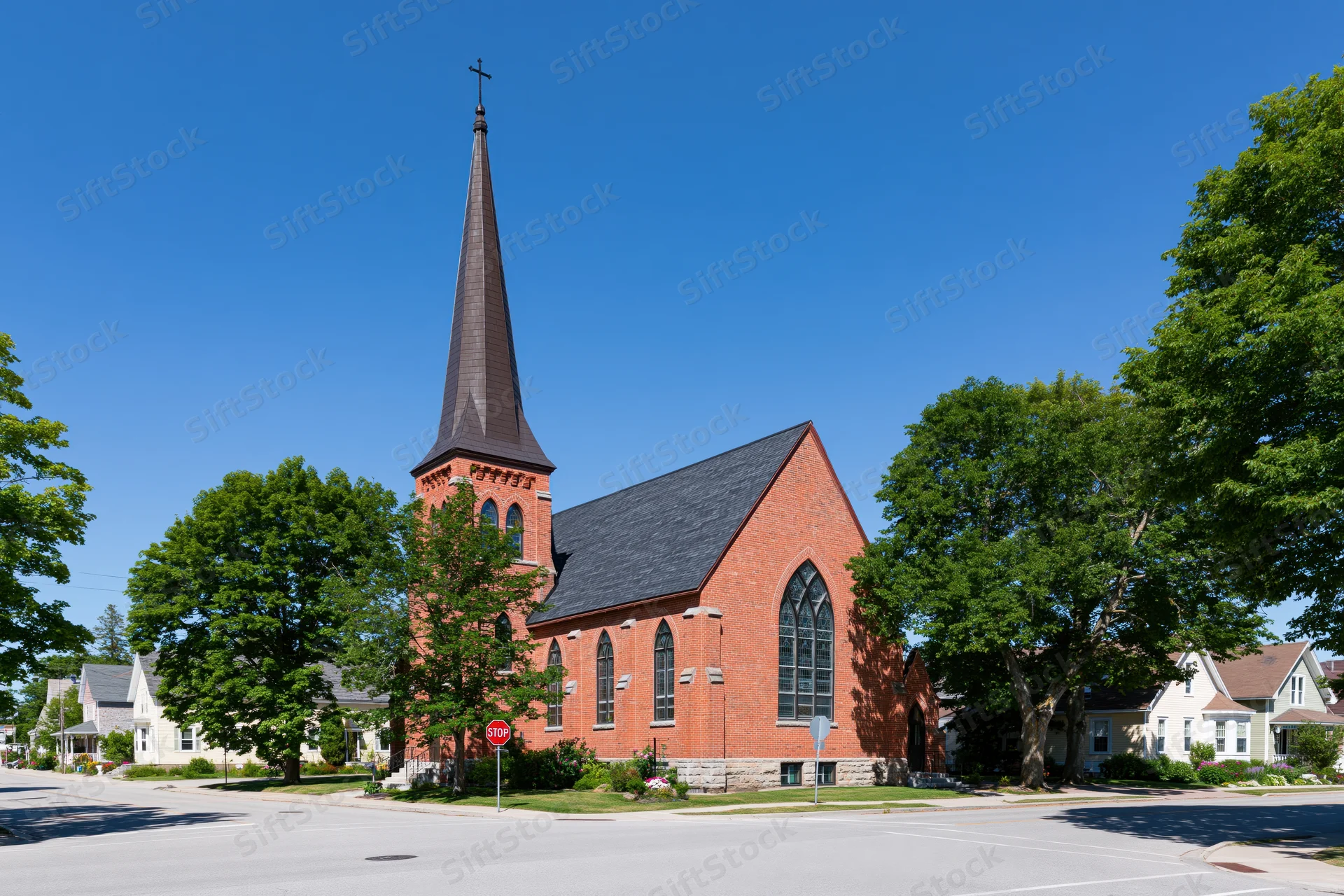 Red-brick gothicrevival church with tall dark steeple and cross at street corner in suburban neighborhood