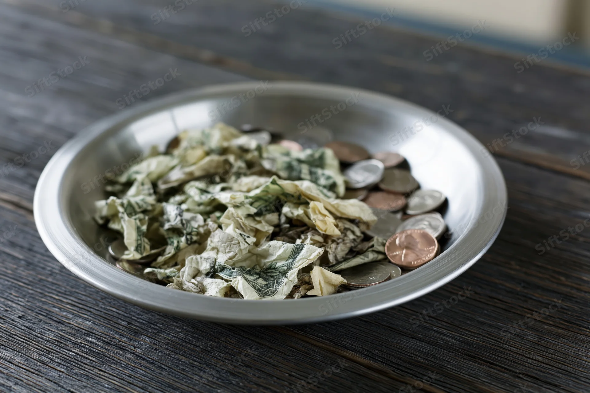 Silver offering plate filled with crumpled us dollar bills and coins on dark wooden surface