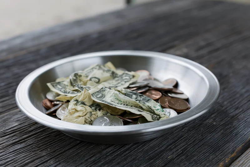 Silver offering plate filled with crumpled us dollar bills and coins on dark wooden table