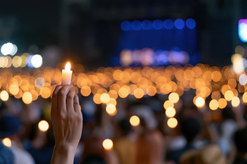 Hand holding lit candle raised high in a nighttime outdoor vigil with blurred crowd and warm bokeh lights in background