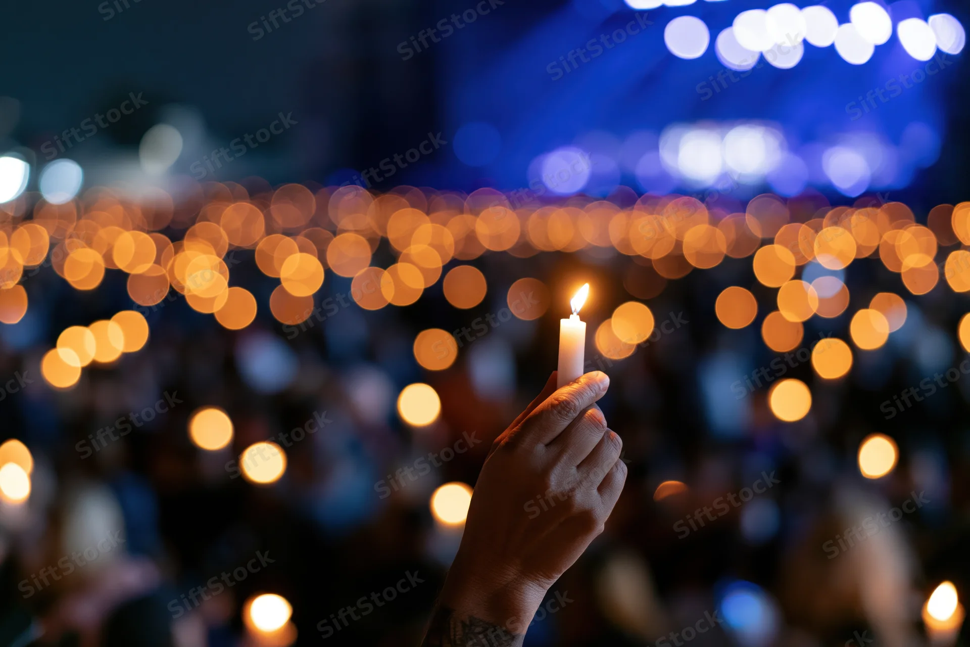 Hand holding lit candle raised high at night during outdoor vigil with blurred lights in background