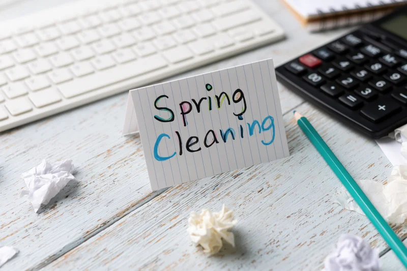 Handwritten spring cleaning note on a cluttered office desk with keyboard, calculator, and crumpled paper