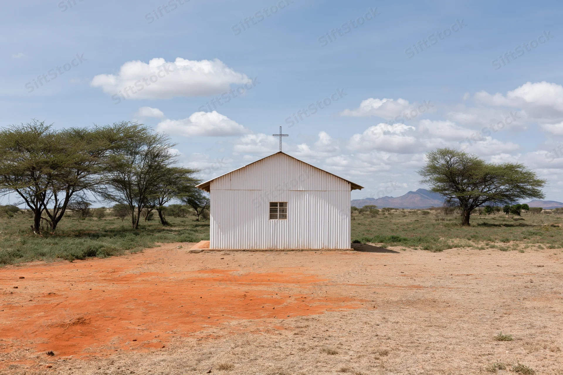 Small white metal church with cross on roof in dry savanna landscape with acacia trees and distant mountains under blue sky with clouds