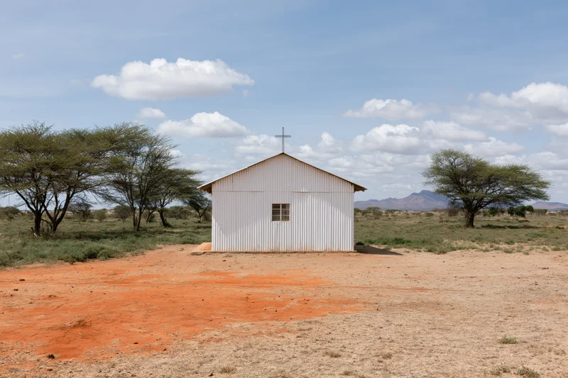 Small white metal church with cross on roof in dry savanna landscape with acacia trees and distant mountains under blue sky with clouds