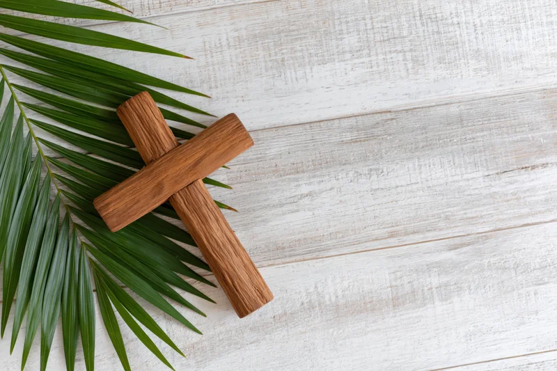 Wooden cross resting beside green palm leaf on white wooden surface