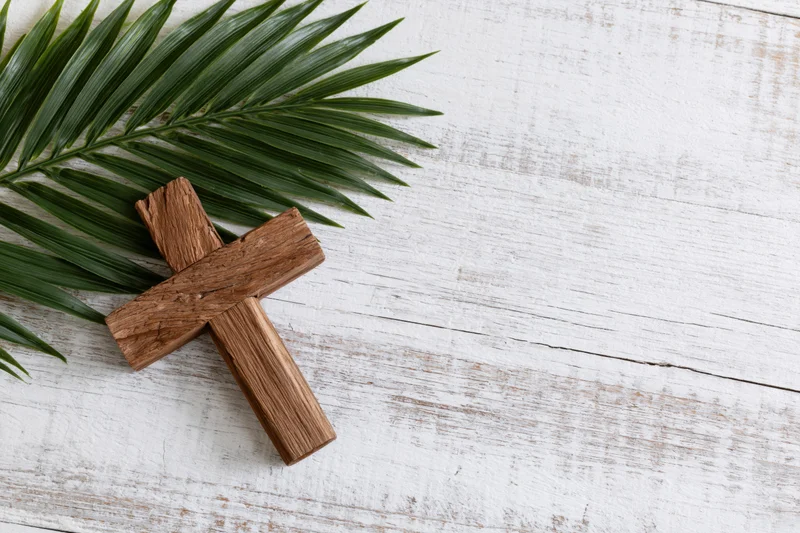 Wooden cross lying beside green palm leaf on white rustic wood surface