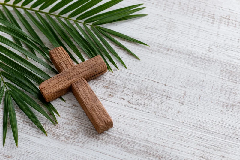 Wooden cross lying flat beside a green palm leaf on a white wooden surface