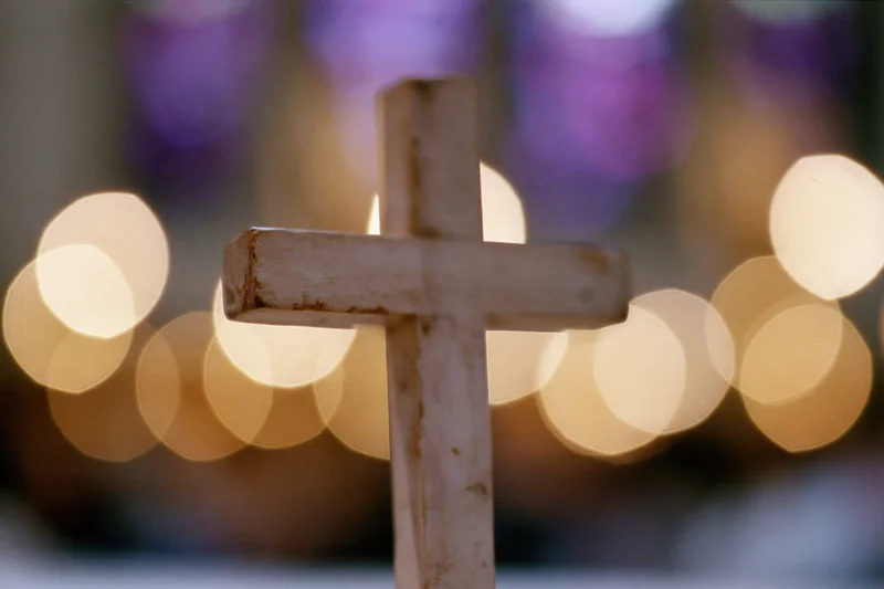 Small worn wooden cross standing upright with blurred warm lights in background