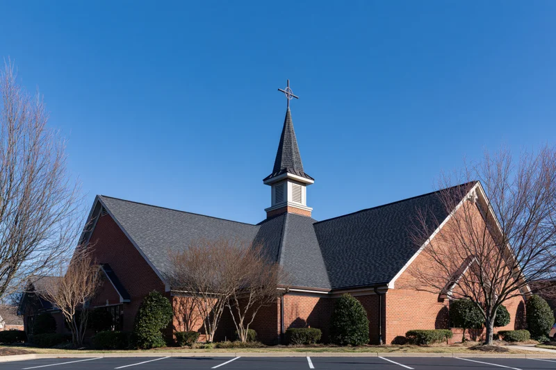 Brick christian church with central steeple and cross under clear blue sky in suburban setting