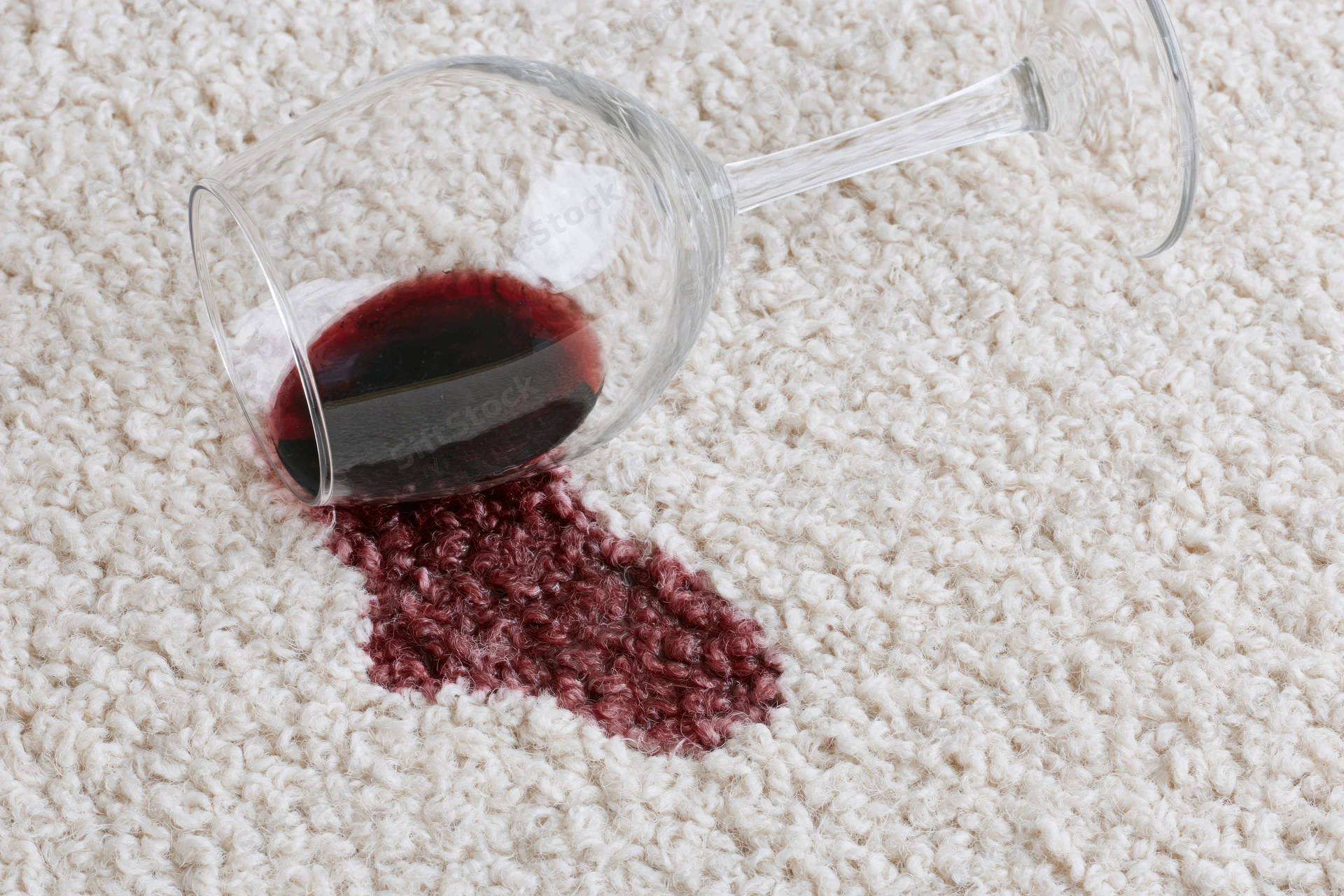 Tipped-over wine glass spilling red liquid onto cream carpet