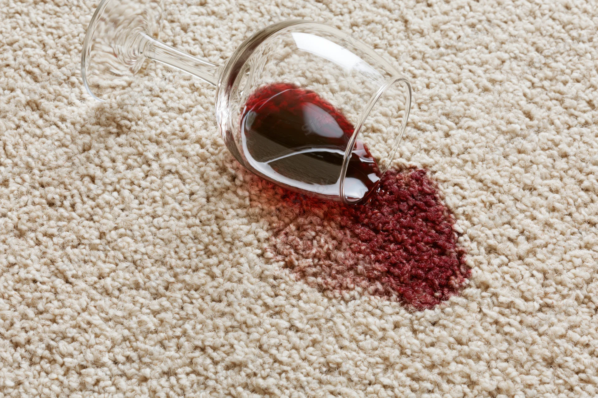 Tipped wine glass spilling red liquid onto beige carpet