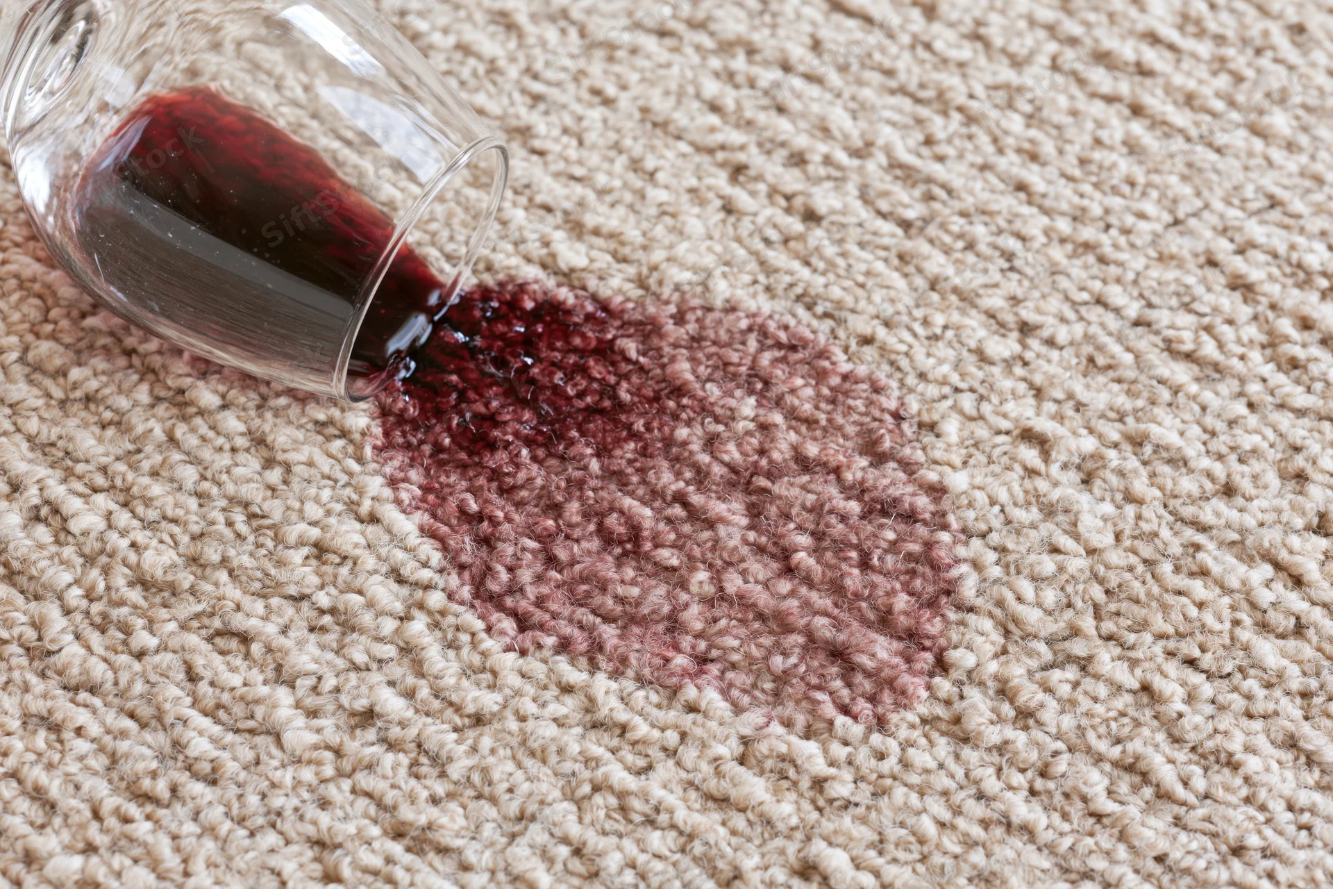 Tipped wine glass spilling red liquid onto beige carpet