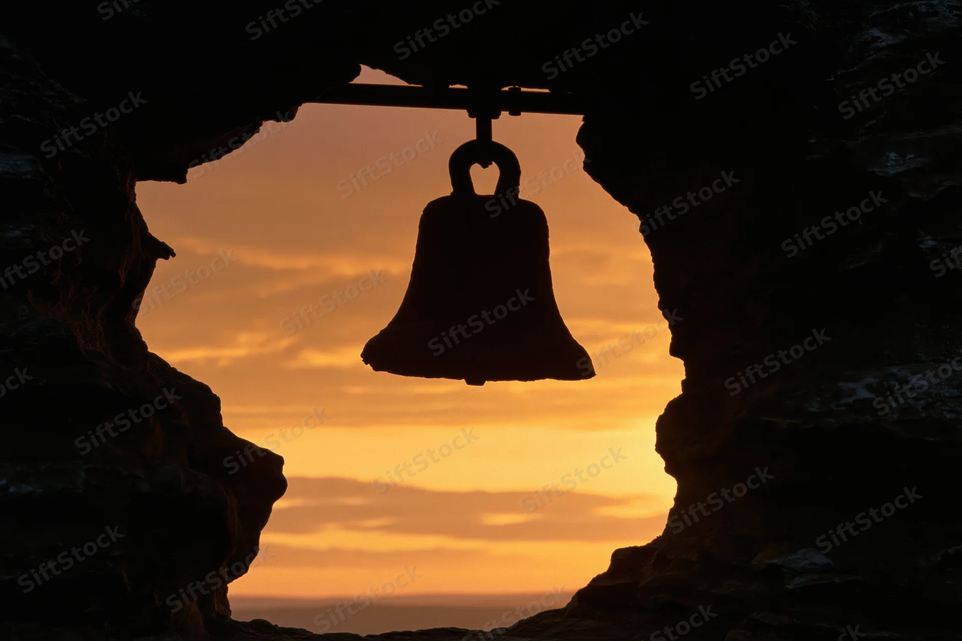 Silhouette of a weathered bronze bell hanging from a rod inside a cave at sunset overlooking the ocean