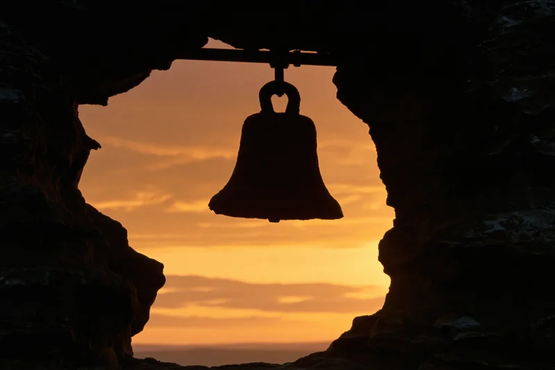 Silhouette of a weathered bronze bell hanging from a rod inside a cave at sunset overlooking the ocean