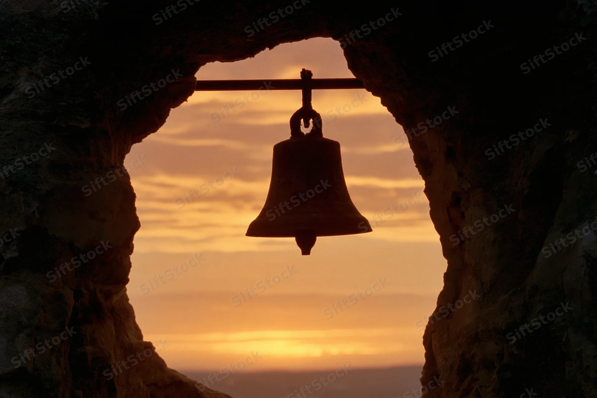 Silhouette of a weathered bronze bell hanging from a rod inside a stone arch at sunset