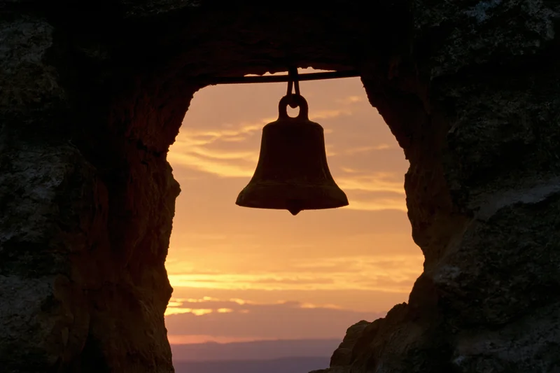 Weathered bronze bell hanging from a rod inside a stone arch at sunset