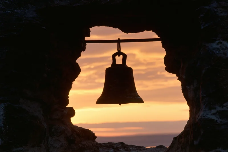 Weathered bronze bell hanging in stone cave opening at sunset