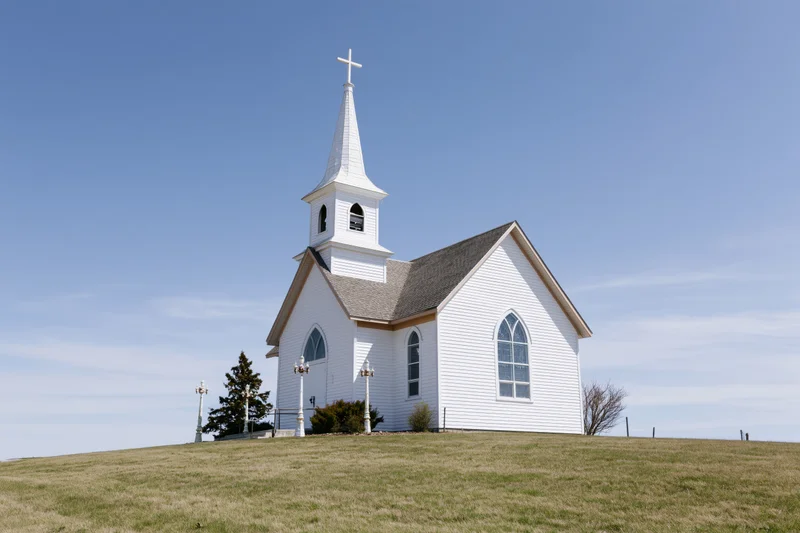 White clapboard church with steeple and cross on a grassy hill under blue sky