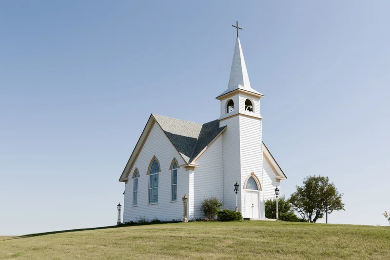 White clapboard church with tall steeple and cross on grassy hill under clear blue sky
