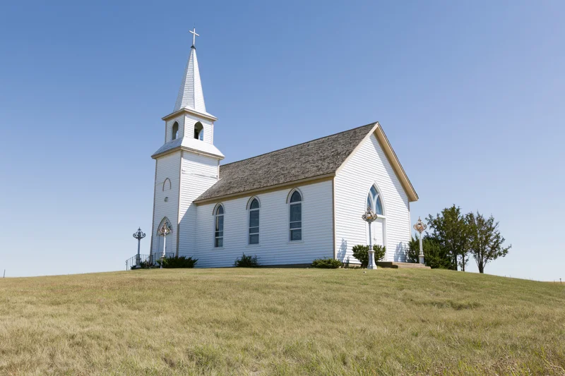 White clapboard country church with tall steeple and cross on grassy hill under clear blue sky