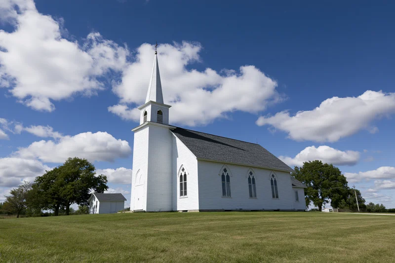 White wooden country church with tall pointed steeple standing on grassy field under blue sky with clouds