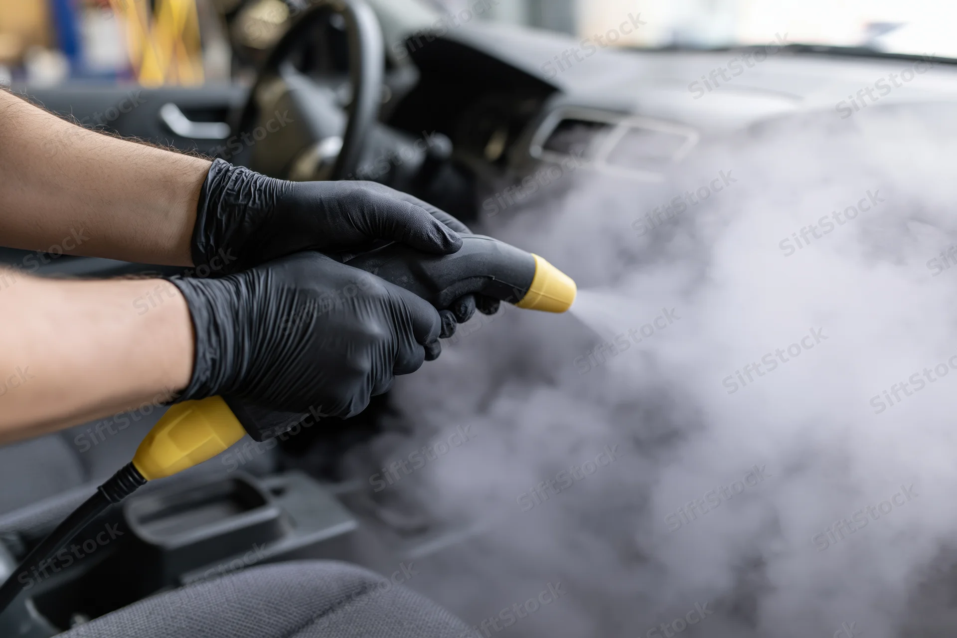 Hands in black gloves using yellow-tipped steam cleaner on car interior