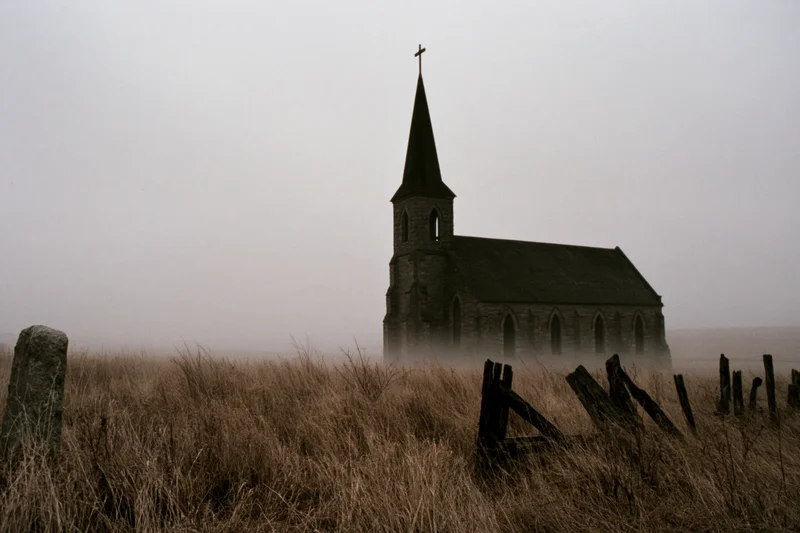 Abandoned gothic stone church with tall steeple and cross in foggy rural field