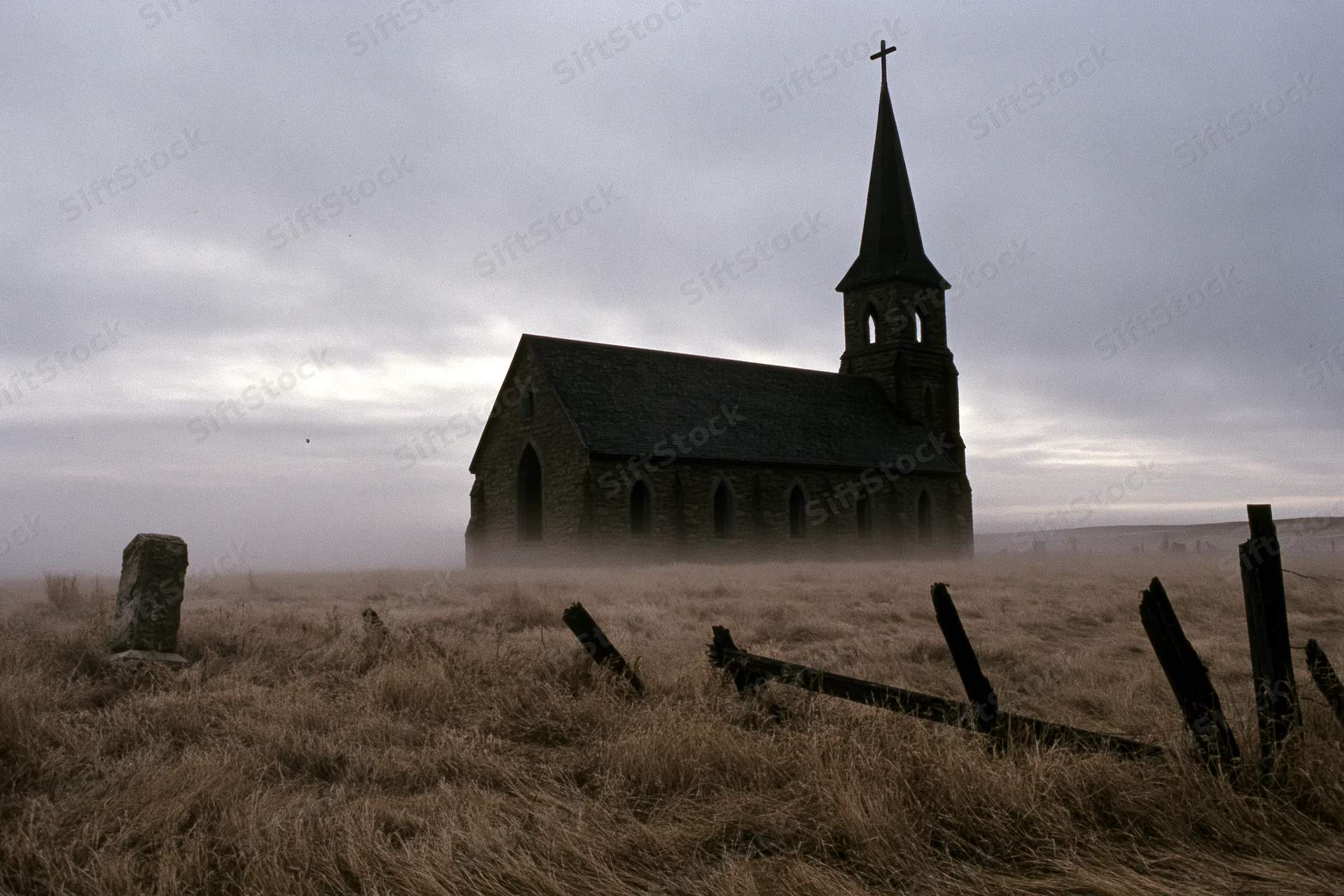 Abandoned gothic stone church with tall steeple and cross in foggy rural field