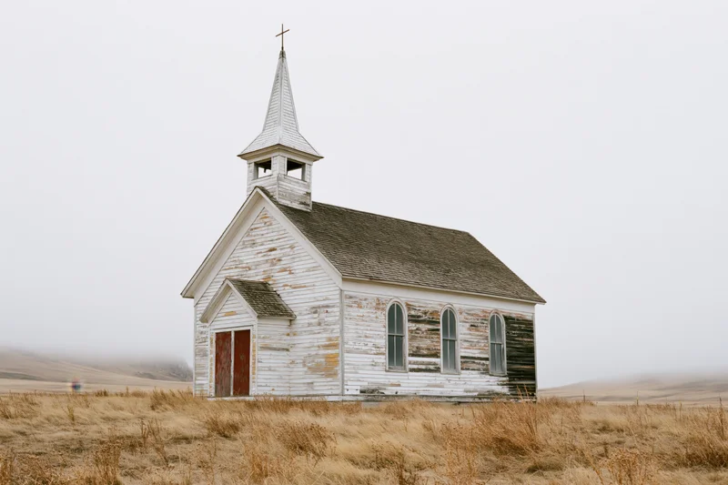 Abandoned white clapboard church with pointed steeple standing in a foggy rural field