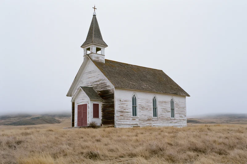 Abandoned white clapboard church with pointed steeple standing in a grassy field under overcast sky